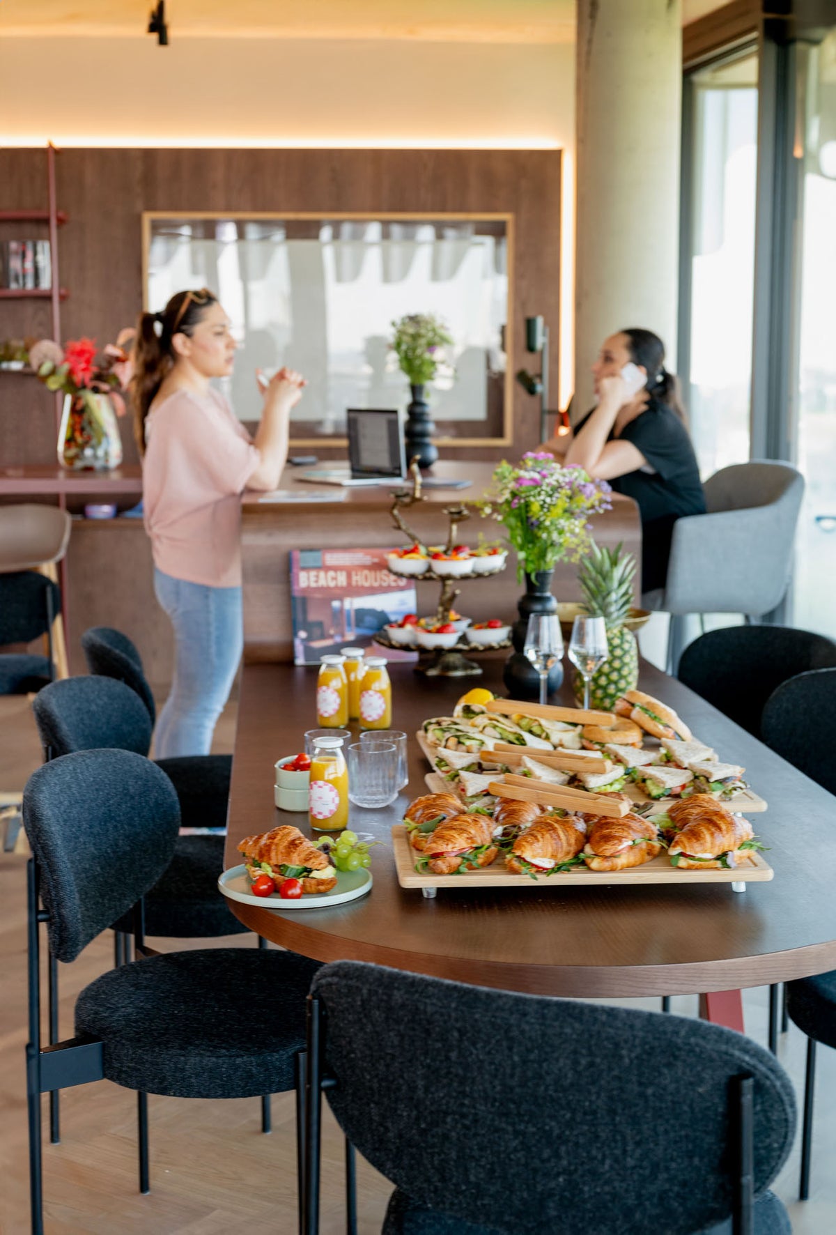 Business meeting table, catering on top, two people blurry in the background, one on the phone, other one talking. Catering includes orange juices, croissants, sandwiches, wraps bagels and fruits. 