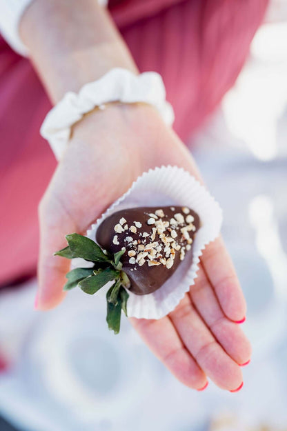 Nahaufnahme einer Hand, die eine frische Erdbeere in dunkler Schokolade mit gehackten Nüssen präsentiert. Die Schokobeere liegt in einer weißen Papierhülle, perfekt als edler Snack für besondere Momente oder Feierlichkeiten.