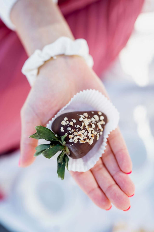 Nahaufnahme einer Hand, die eine frische Erdbeere in dunkler Schokolade mit gehackten Nüssen präsentiert. Die Schokobeere liegt in einer weißen Papierhülle, perfekt als edler Snack für besondere Momente oder Feierlichkeiten.