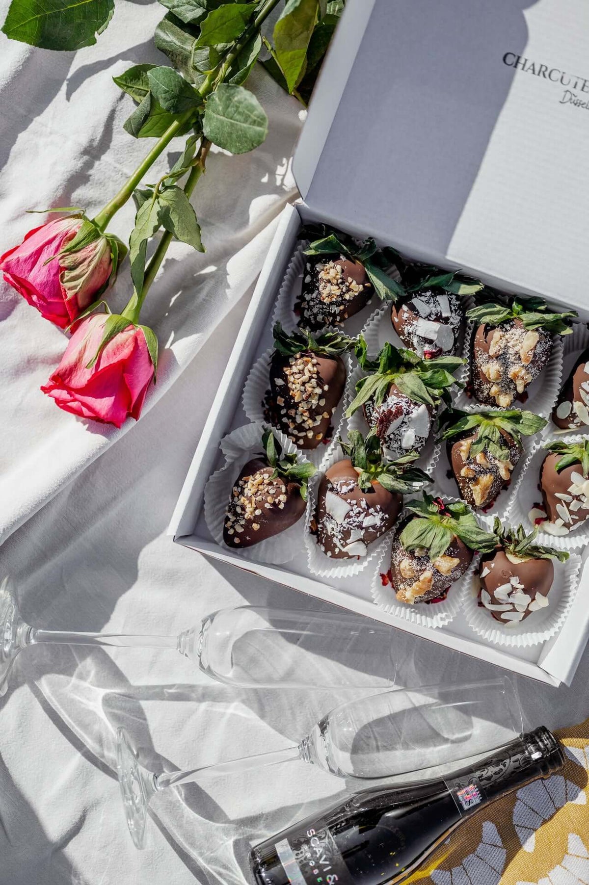 Schokobeeren-Box mit frischen Erdbeeren, umhüllt von dunkler Schokolade und verziert mit Nüssen und Kokosraspeln. Präsentiert auf einem Tisch mit Sektflasche, Gläsern und Rosenblättern, mit Blick auf eine malerische Wasserlandschaft – ideal für besondere Anlässe und romantische Momente.